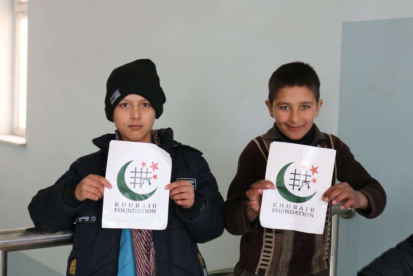 Two young boys holding papers with the Khubaib Foundation logo.